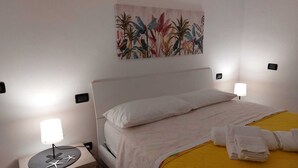 En snyggt bäddad säng med vita sängkläder, två sänglampor, en tavla med tropiska växter som hänger på väggen och ett litet bord med en sjöstjärnedekoration och en lampa.