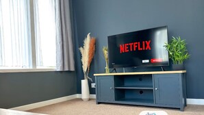 A television on a stand with 'NETFLIX' displayed, a vase with dried plants, and a window with curtains. - Large House | Convention Centre | 20% Discount (Harrogate)