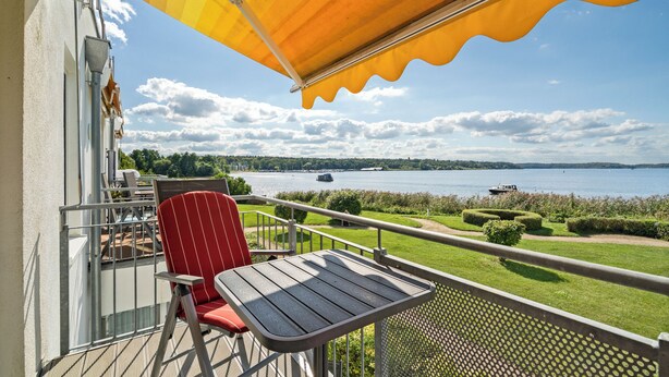 A balcony with a table and a red chair, overlooking a lake with boats and a distant shoreline. - Vacation apartment "Seepferdchen" with a fantastic view of the Müritz (Waren (Müritz))