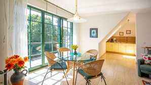 A dining area with a glass table and wicker chairs, a large window with green frames, and a kitchen in the background. - "Kranichruh" Vacation Rental in Sembzin, Nestled in the Heart of Mecklenburg's Natural Beauty (Klink)