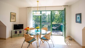 A dining area with a glass table and wicker chairs, a television on a glass cabinet, and a view of greenery through a green door. - "Kranichruh" Vacation Rental in Sembzin, Nestled in the Heart of Mecklenburg's Natural Beauty (Klink)