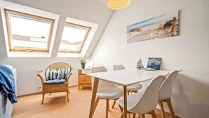 A dining area with a white table and chairs, a wicker chair with a cushion, a potted plant, and a beach-themed picture on the wall. - Vacation apartment "Studio" by the lake in Waren (Müritz) (Waren (Müritz))