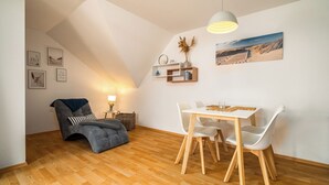 A loft-style room with a dining table, white chairs, a gray sofa, wooden flooring, a ceiling lamp, wall art, and a small shelf with decorative items. - Vacation apartment "MüritzLounge" in Waren (Müritz) (Waren (Müritz))