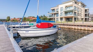 Ein modernes Gebäude mit Balkonen und großen Fenstern, eine Anlegestelle mit einem Segelboot und ruhigem Gewässer.