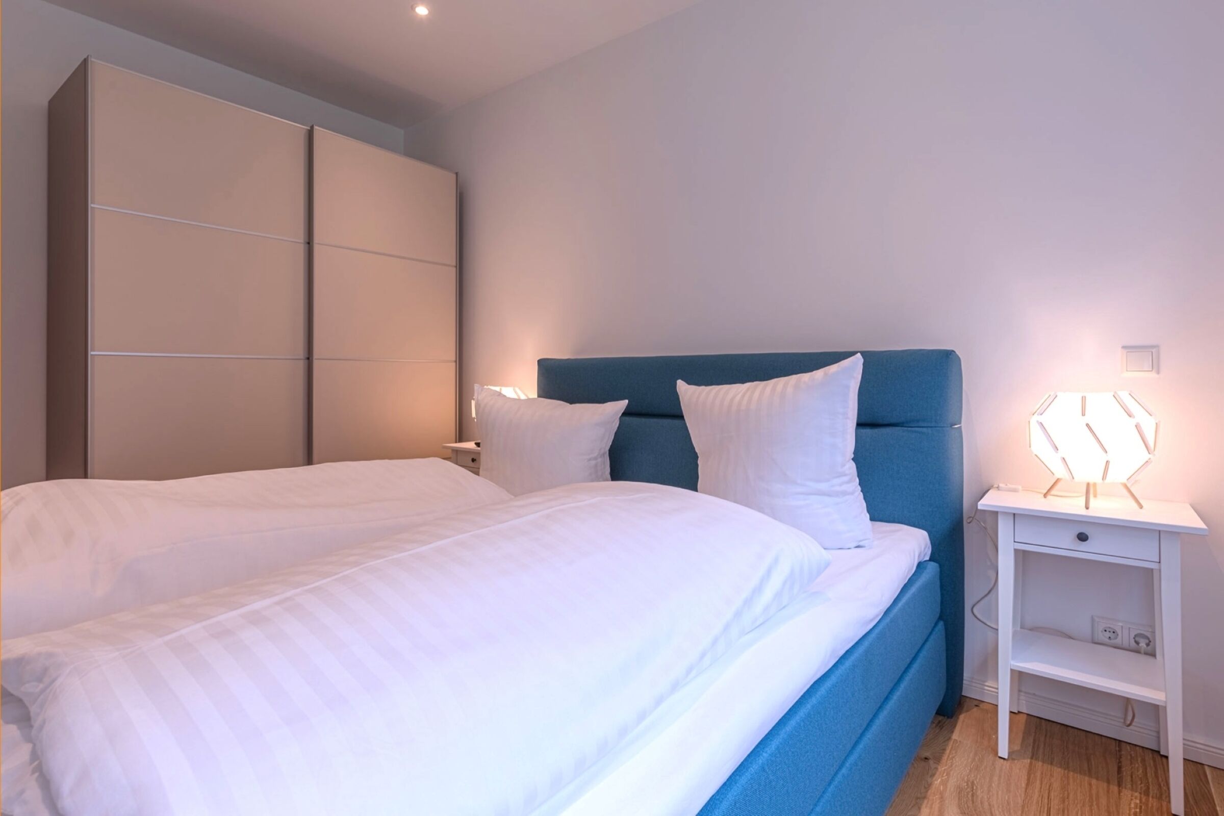 Ein Schlafzimmer mit einem großen Bett, einem blauen Kopfteil, weißer Bettwäsche, einem weißen Nachttisch mit Lampe und einem Kleiderschrank.