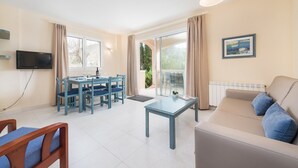 A modern living room with a sofa, a dining table with chairs, a TV, and a large window with curtains. - Oro Del Mar Iv Apartamento O2 - Apartment (Canyamel - Capdepera, Illes Balears)