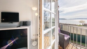 Een balkon met een televisie, lamp en rieten stoel met uitzicht op een waterpartij.