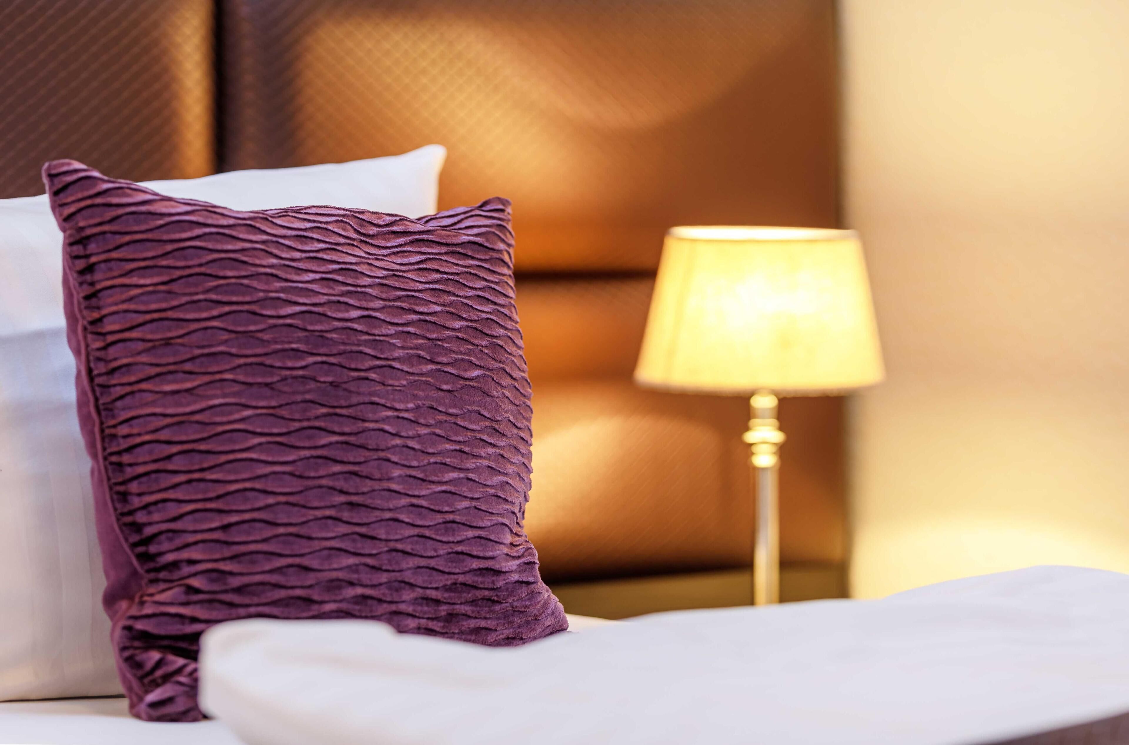 A neatly made bed with a purple textured pillow and a white pillow, a bedside lamp, and a headboard.