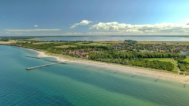 Luftaufnahme einer Küstenlandschaft mit Strand, einem Steg und einer Wohngegend mit Häusern und Grünflächen.