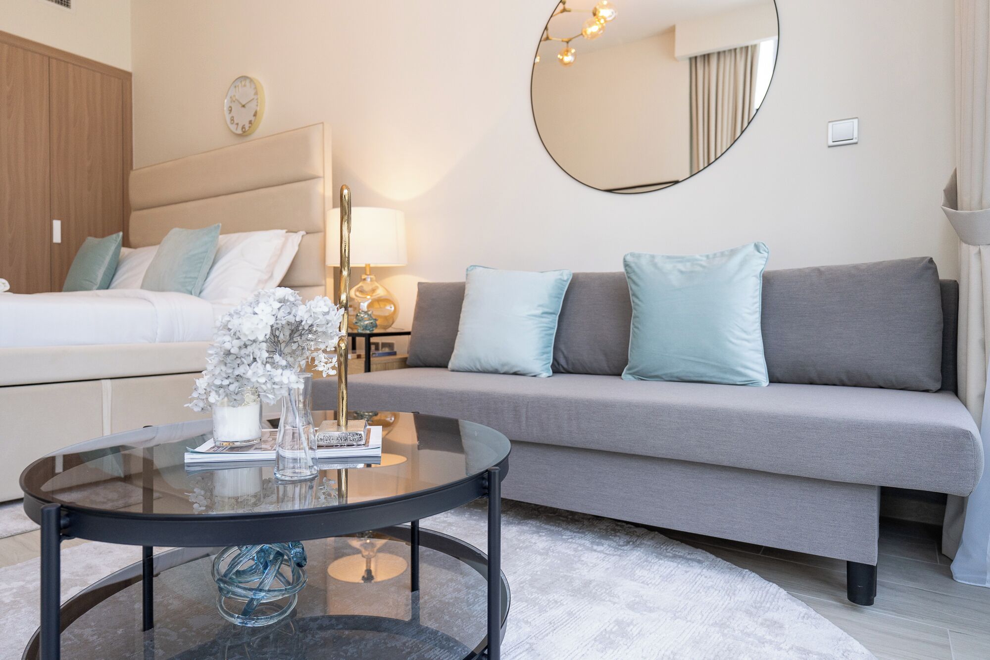 A modern bedroom with a bed, a grey sofa, a round mirror, and a glass coffee table with decorative items.