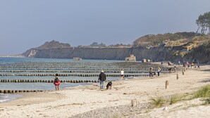 A beach with a rocky cliff in the background, a wooden jetty, and people walking along the shore.