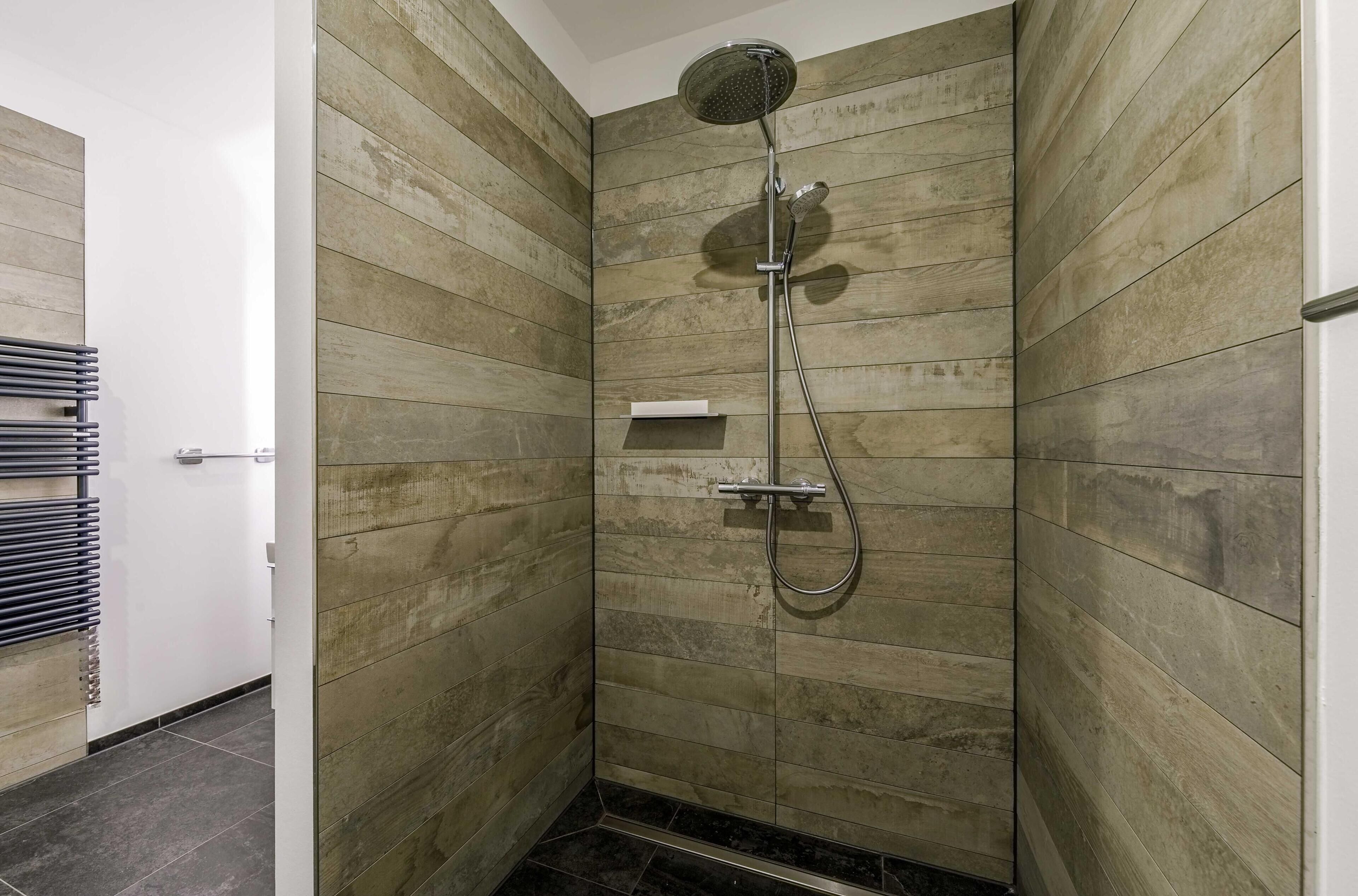 Eine Dusche mit Handbrause und Wandarmatur, eingebettet in eine Ecke mit Holzwandverkleidung und einem kleinen Ablagebrett.