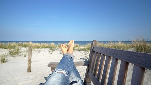 A wooden bench with a person sitting on it, facing a beach with sand and grass.
