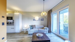 A modern living room with a sofa, a coffee table, and a dining table. There is a kitchen area with white cabinets and a microwave. The room has large windows with curtains and a view of the outdoors. - Vacation apartment "Große Stranddüne" in Wustrow on the Baltic Sea (Wustrow)