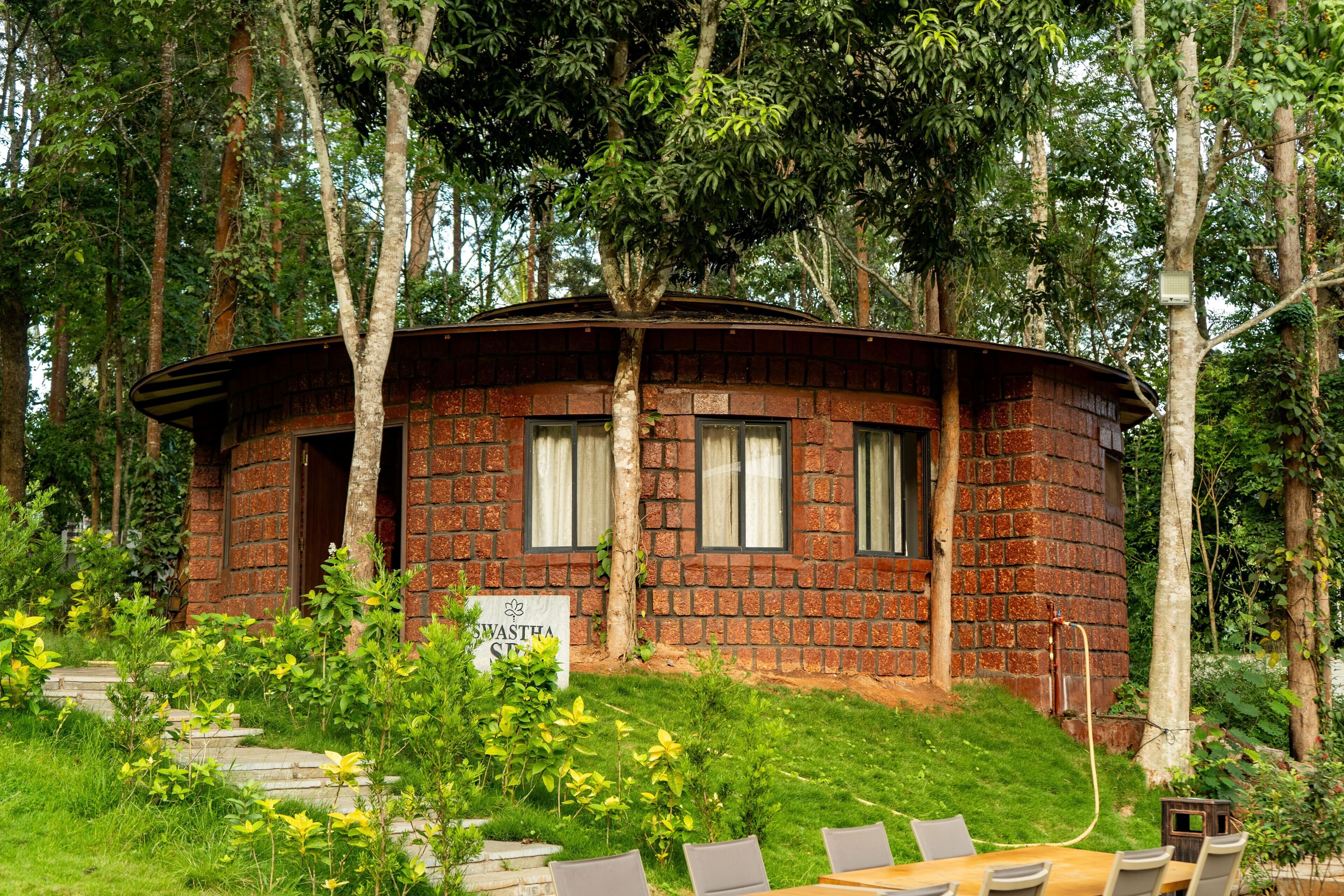 A circular brick building with a sign reading 'Swasta Spa' surrounded by lush greenery and a wooden table with chairs.