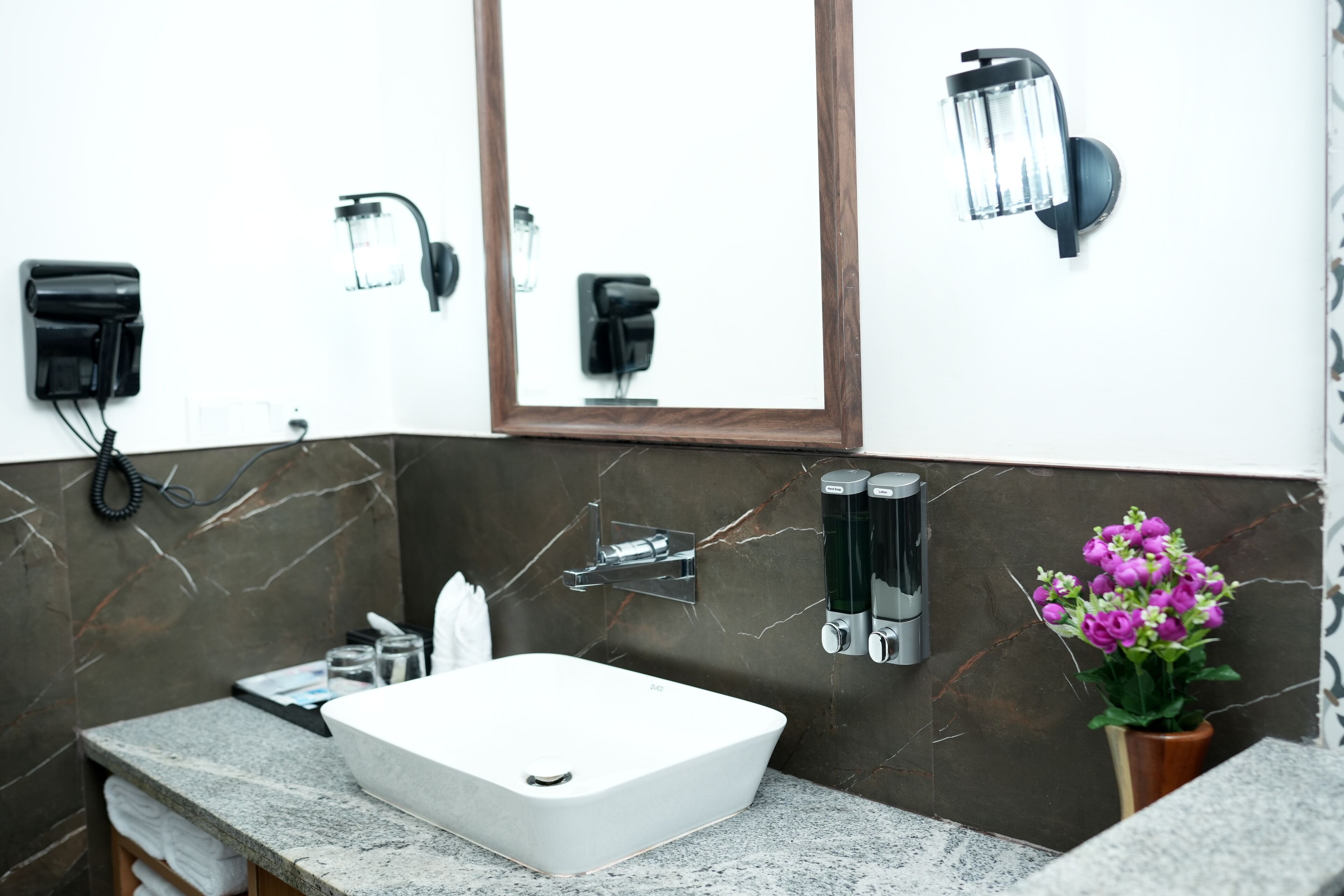 Un baño moderno con un lavabo blanco, salpicadero de mármol y un espejo encima. Hay dos dispensadores de jabón montados en la pared y una pequeña planta en maceta con flores moradas.