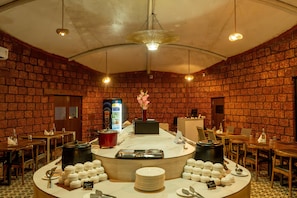 A restaurant with brick walls, a central counter with food items, and tables with chairs. - TGI Redolent Resort & Spa (Somvarpet)