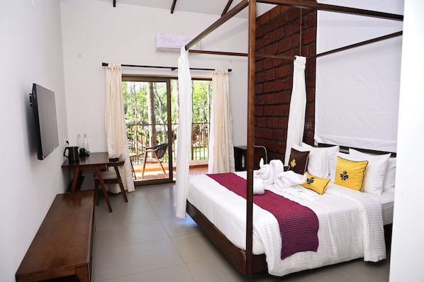 A four-poster bed with white bedding and yellow pillows, a wooden desk, a television, and a balcony with a chair. - TGI Redolent Resort & Spa (Somvarpet)