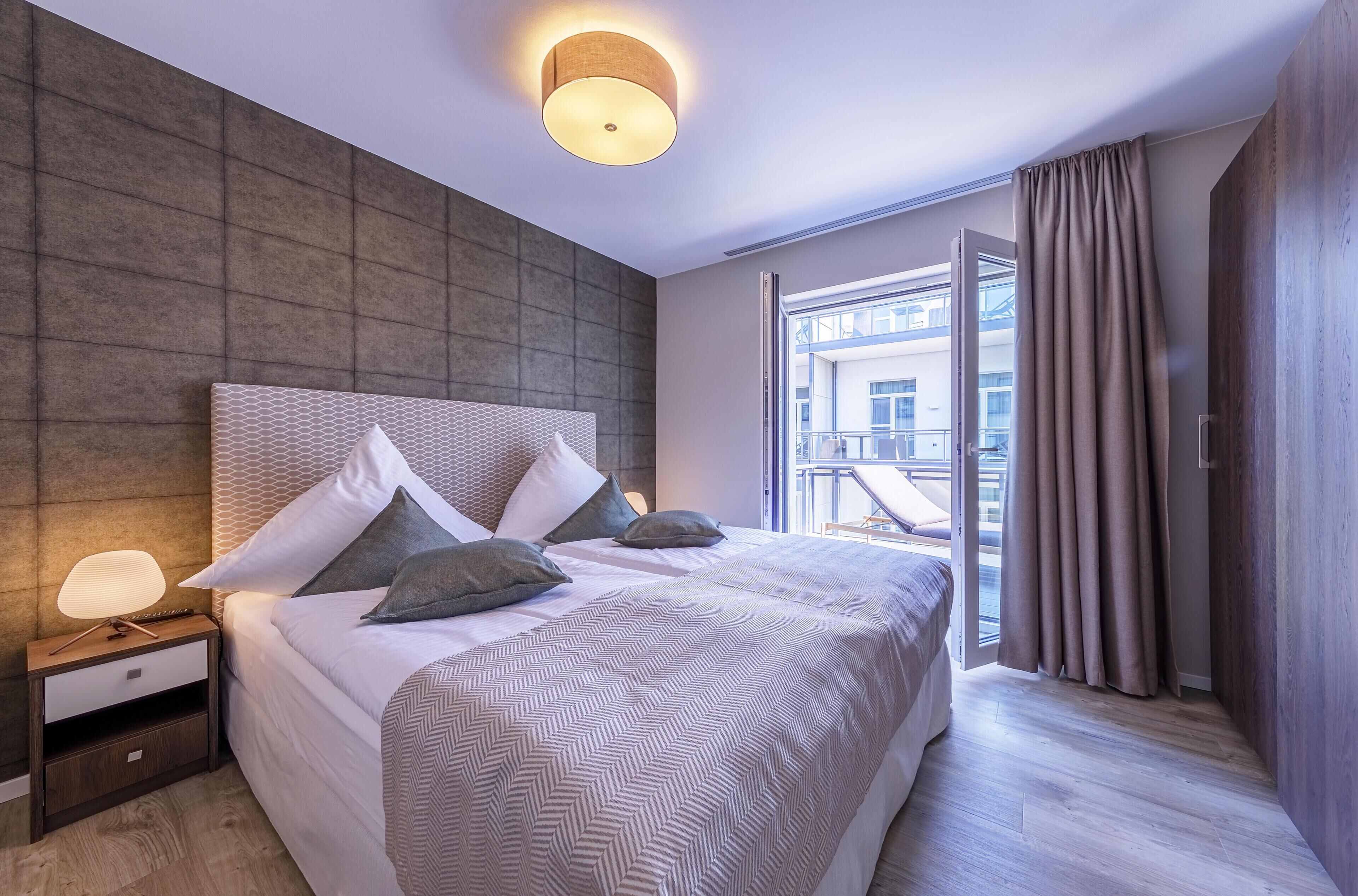 Ein modernes Hotelzimmer mit einem großen Bett, einem Nachttisch, einer Wandleuchte und Blick auf die Stadt durch große Fenster.
