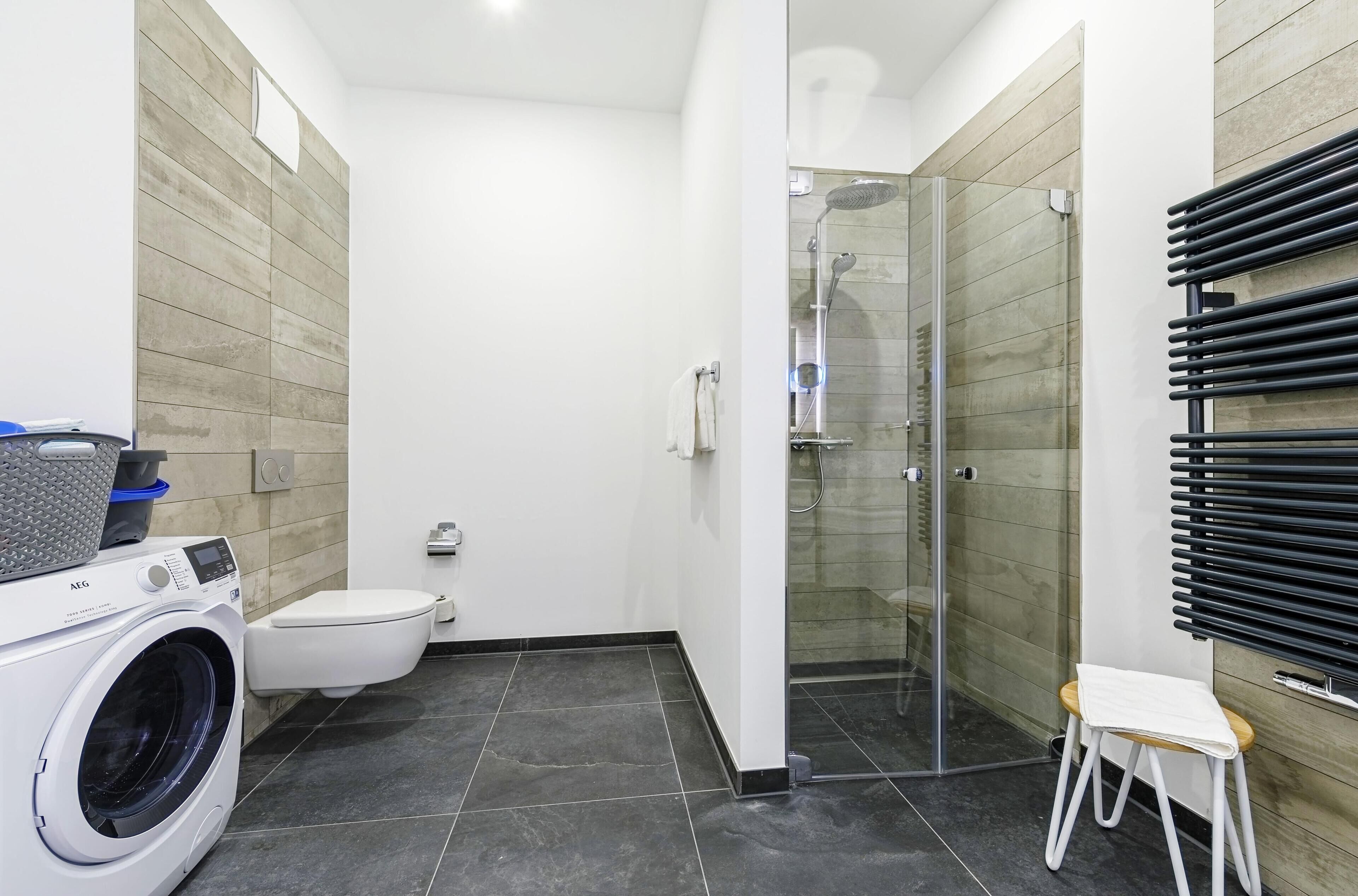 Et moderne badeværelse med vaskemaskine, toilet, brusekabine i glas og en lille skammel.