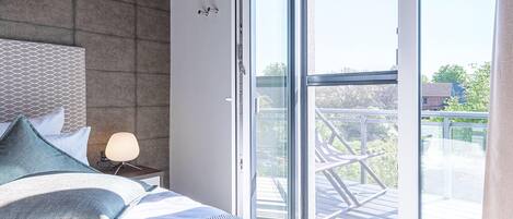 A bedroom with a bed, pillows, a bedside table with a lamp, and a large sliding glass door leading to an outdoor balcony.
