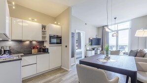 Une cuisine moderne avec des meubles blancs, un four encastré et une machine à café. On y trouve également un coin repas avec une table en bois foncé et des chaises de couleur beige.