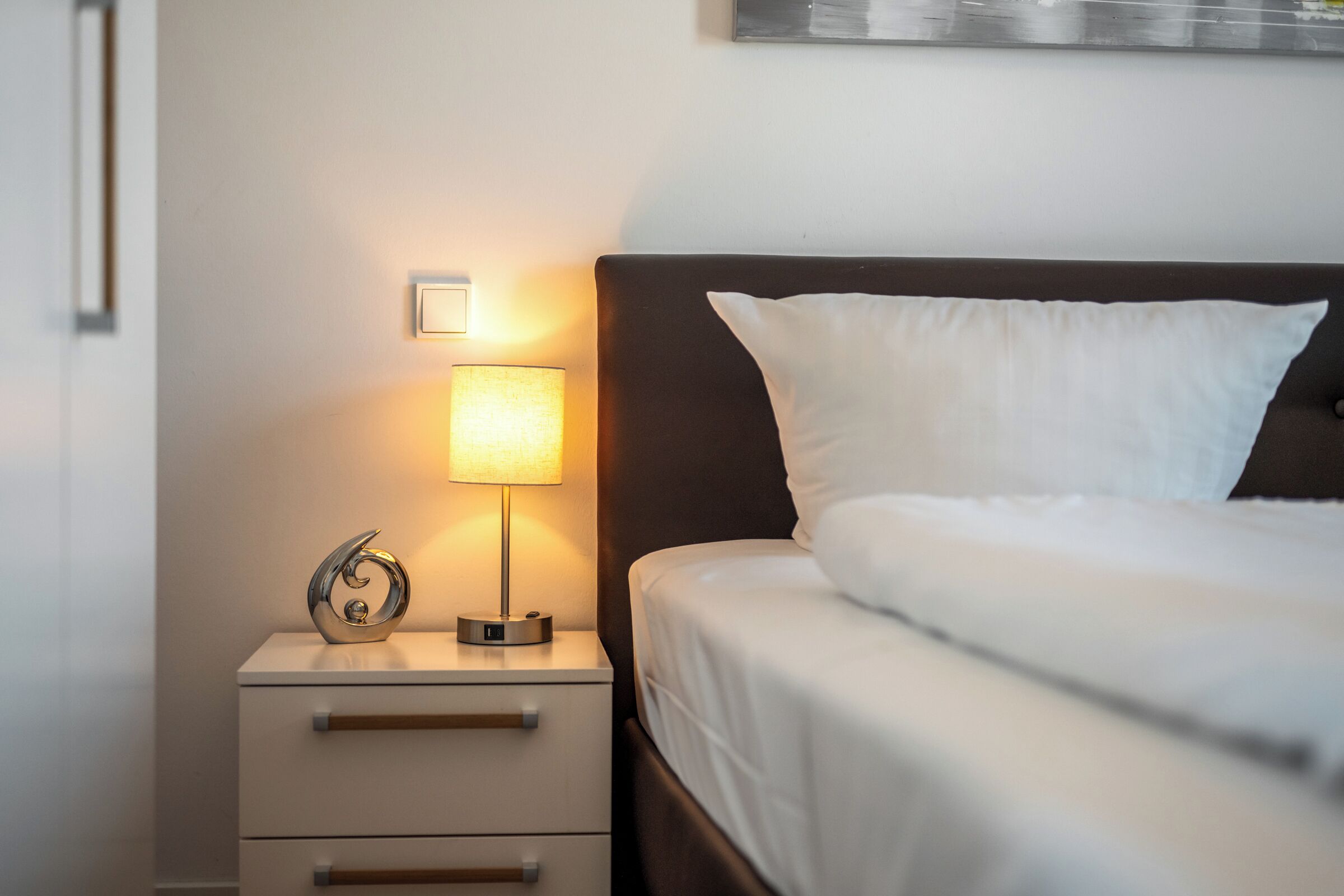 Una cama bien hecha con ropa blanca, una lámpara de noche, un objeto decorativo pequeño y una mesita de noche con cajones.