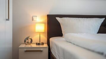 Una cama bien hecha con ropa blanca, una lámpara de noche, un objeto decorativo pequeño y una mesita de noche con cajones.