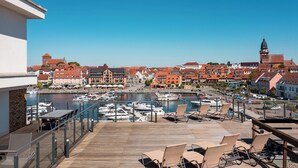 Una terraza en la azotea con suelo de madera, sillones de descanso y vista a un puerto deportivo con barcos y edificios al fondo.