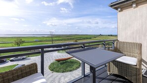 Un balcon avec une table et des meubles en osier, donnant sur un paysage verdoyant et une route où circulent des véhicules.