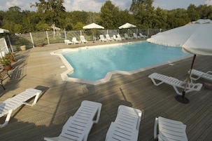 Piscina com espreguiçadeiras brancas e guarda-sóis numa plataforma de madeira, cercada por uma vedação e árvores.