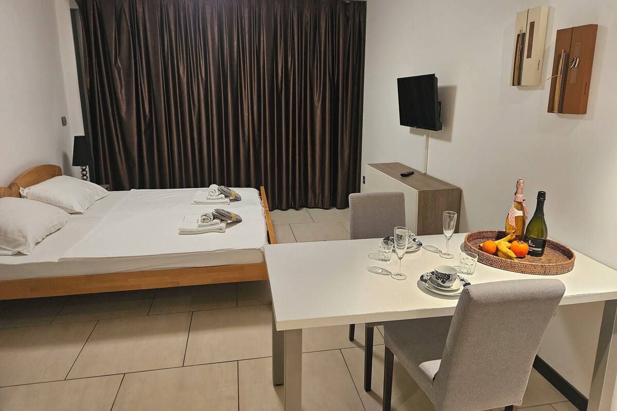 A hotel room with a bed, a desk with a chair, a television, and a table set with a bottle, glasses, and fruit.