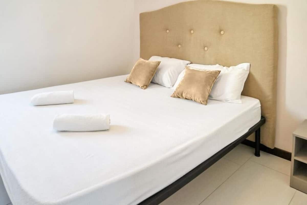A neatly made bed with white linens and two beige pillows, a wooden headboard, and a small bedside table.
