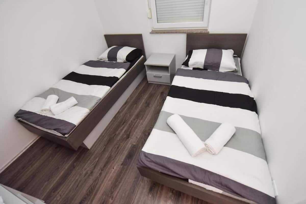 A room with two single beds, each with a wooden frame and striped bedding. There is a small bedside table and a drawer unit between the beds.