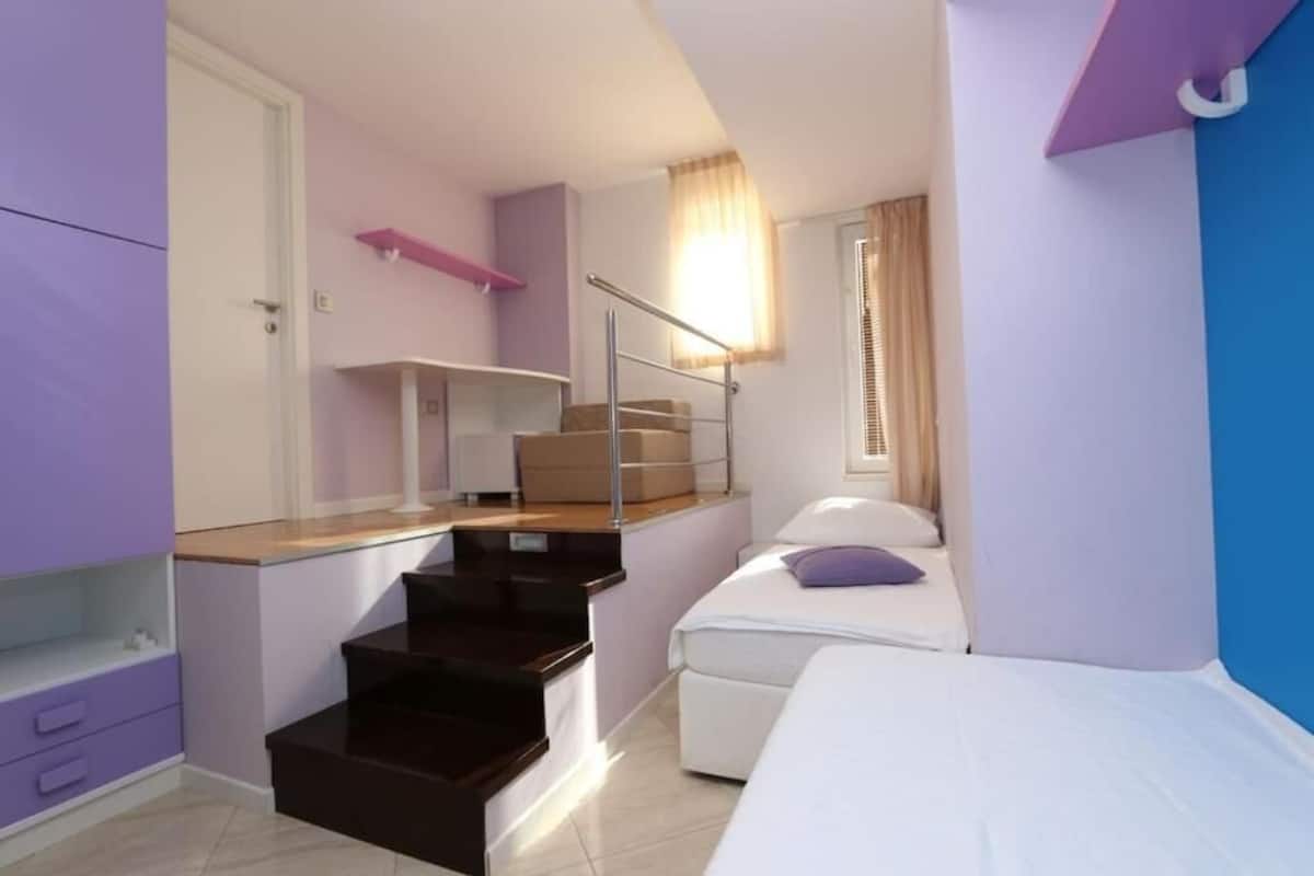 A compact bedroom with a bed, a small desk, and a staircase leading to an upper level.