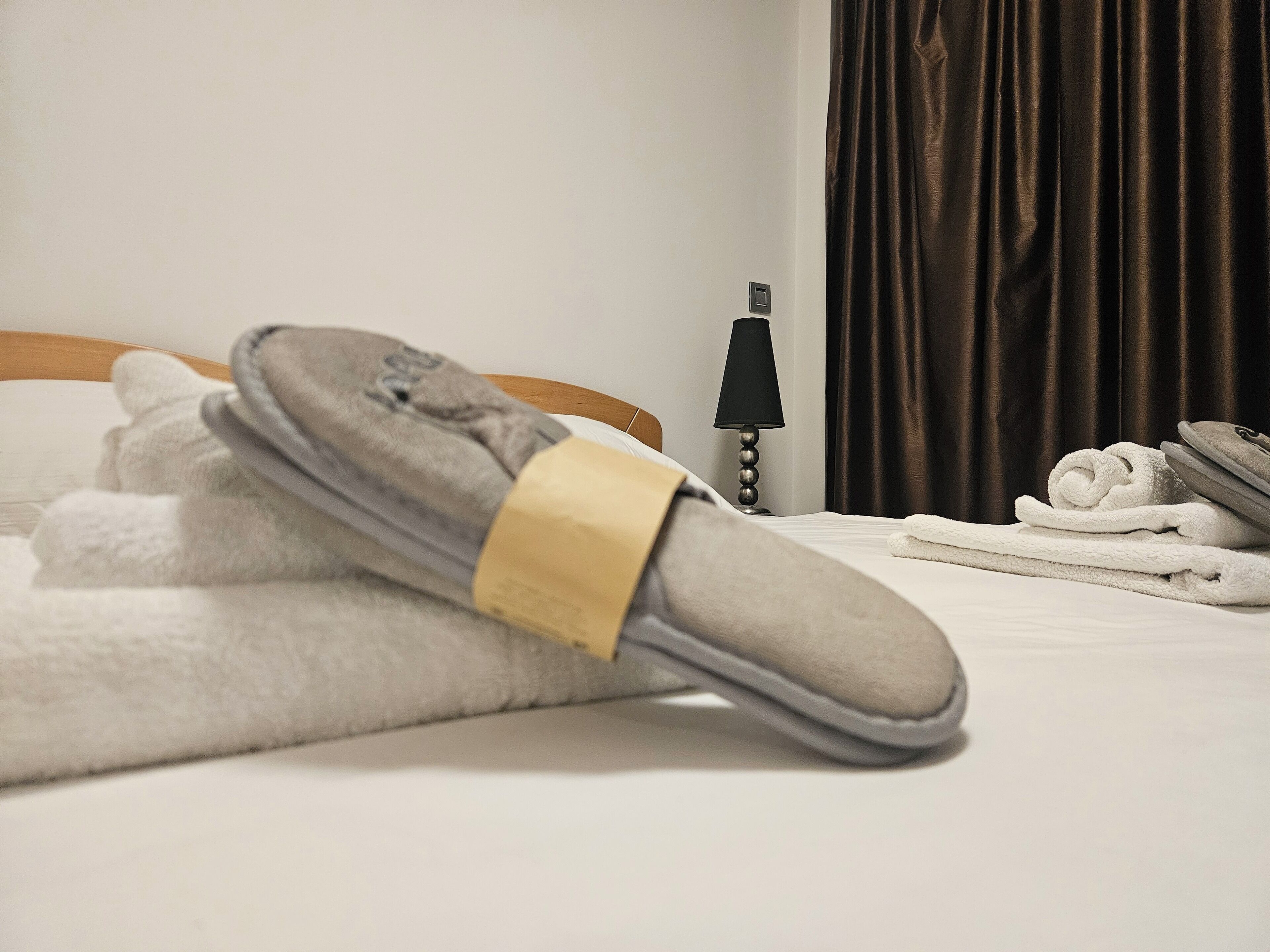 Una cama bien tendida con un par de zapatillas, una lámpara y una toalla enrollada al lado.