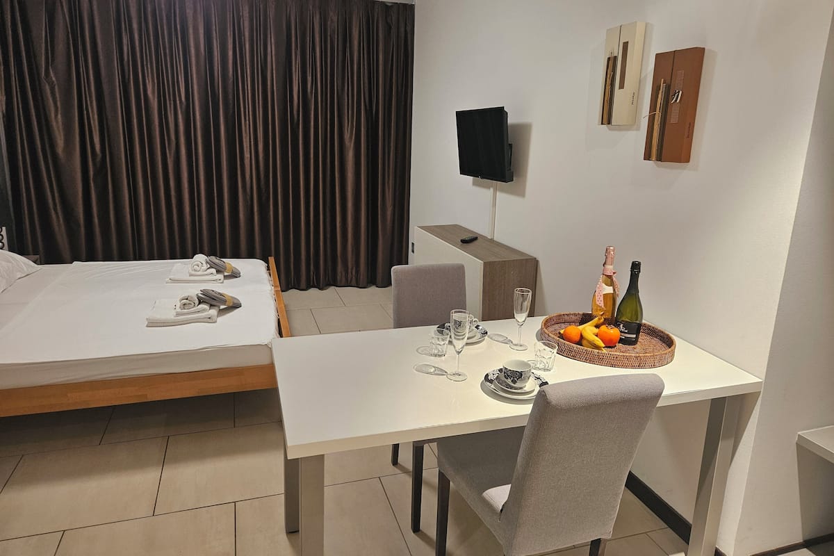A hotel room with a bed, a dining table with a chair, a TV mounted on the wall, and a small table with a bottle and fruits.