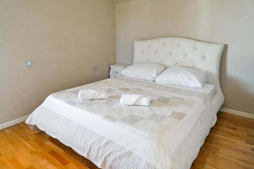 A neatly made bed with a white duvet and pillows, a wooden floor, and a simple nightstand.