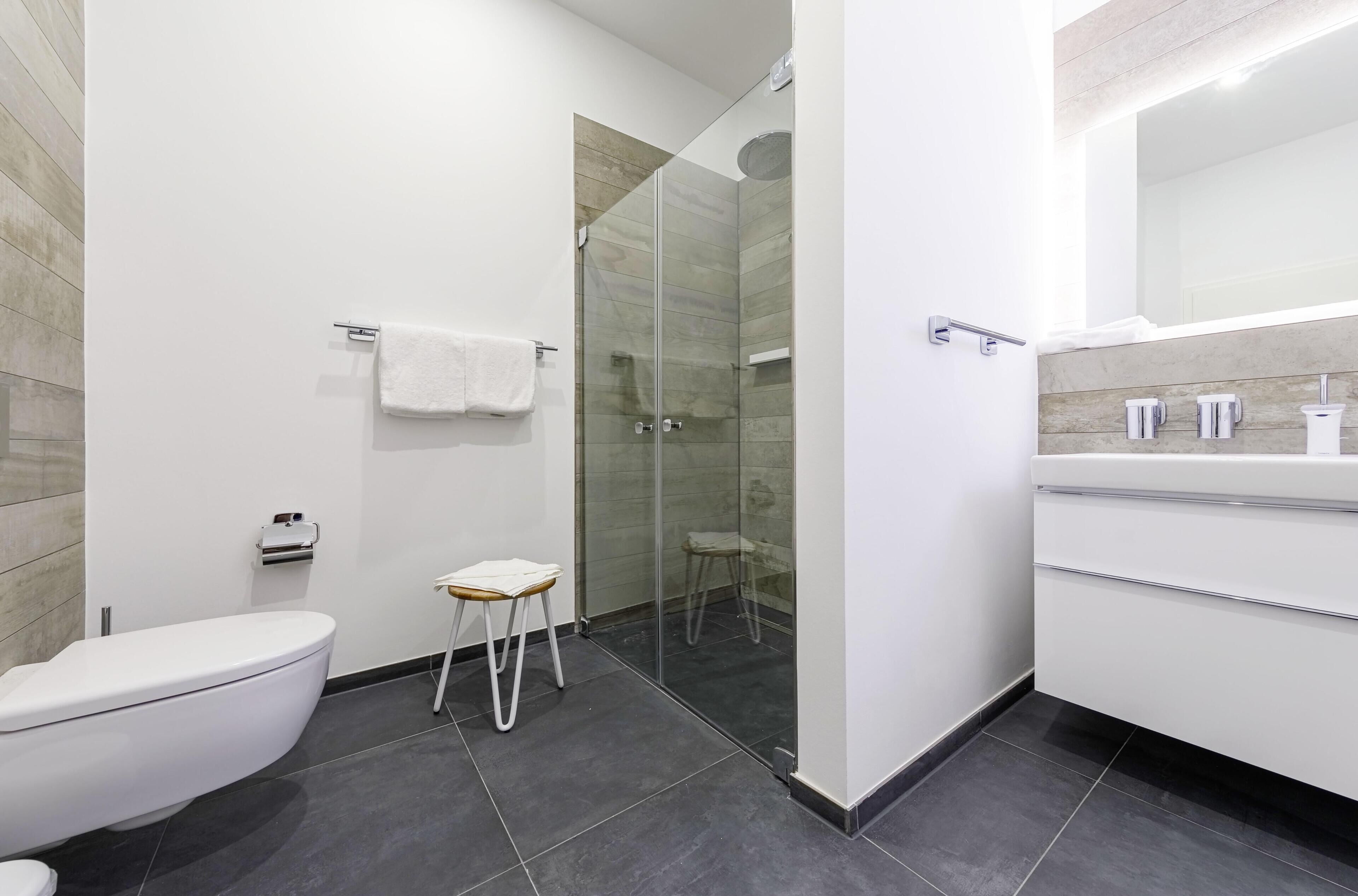 A modern bathroom with a toilet, a glass-enclosed shower, a small stool, and a vanity with a mirror.