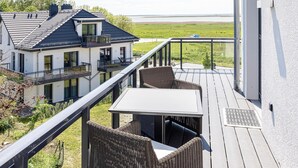 A balcony with wicker furniture and a table, overlooking a grassy area and a body of water. - "Vitus Bering Suite" Vacation Rental with a Balcony on the Baltic Sea (Wustrow)