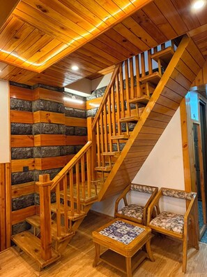 A wooden staircase with a handrail leading to a landing area with two chairs and a small table. - The Uttrayan (Chakrata)