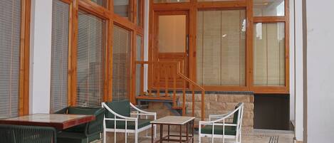 Una habitación con escalera de madera, sillas verdes, una mesa y ventanas grandes con persianas.