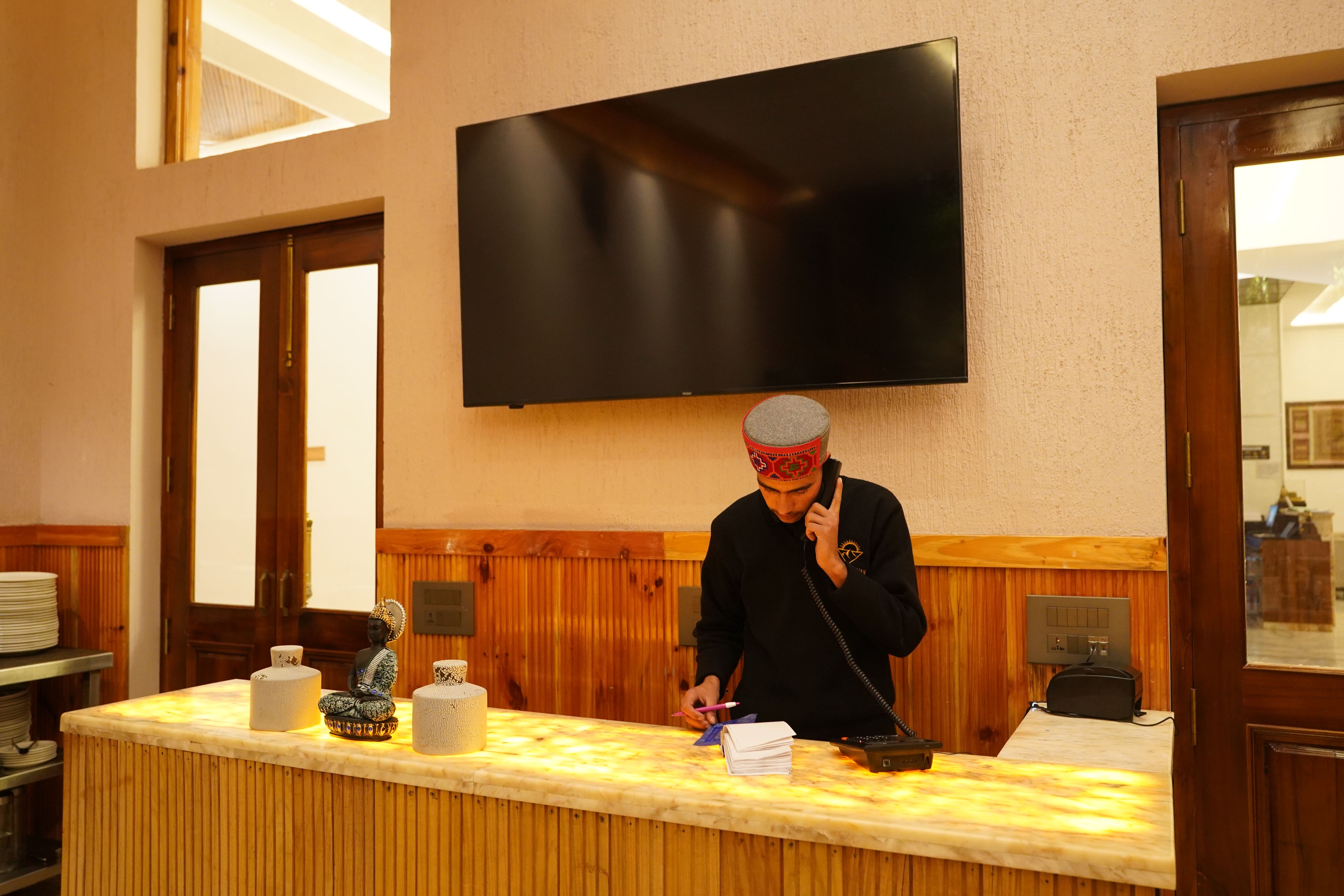 A reception desk with a flat-screen TV mounted on the wall, wooden paneling on the walls, and a counter with various items including a small Buddha statue and a stack of papers.