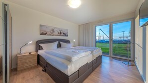 A modern bedroom with a large bed, bedside tables, a painting, and a view of a green landscape through large windows.