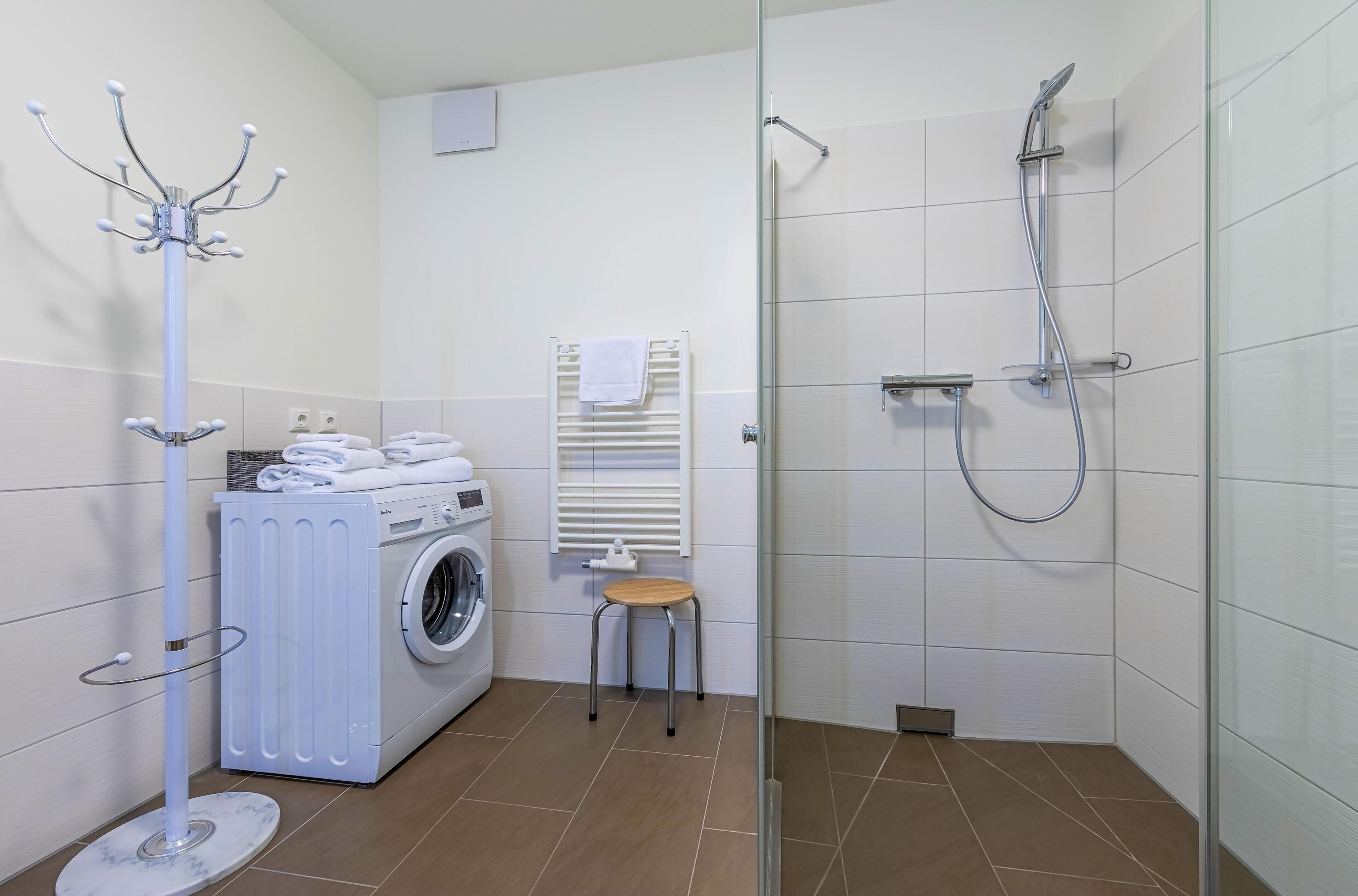 Une salle de bain moderne équipée d’un lave-linge, d’un porte-serviettes, d’une cabine de douche en verre et d’un petit tabouret.