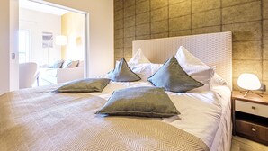 Hotelkamer met een groot bed, meerdere kussens, een nachtkastje met een lamp en uitzicht op een andere kamer door een geopende deur.