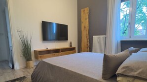 A bedroom with a bed, a TV mounted on the wall, a wooden shelf, and a window with a view. - GuestHost - 65 sqm apartment for 4 people, located on the second floor of a building without an elevator, featuring a balcony. Situated in the Lingotto neighborhood, a convenient and well-served area, close to the Regina Margherita Hospital and just minu (Torino)