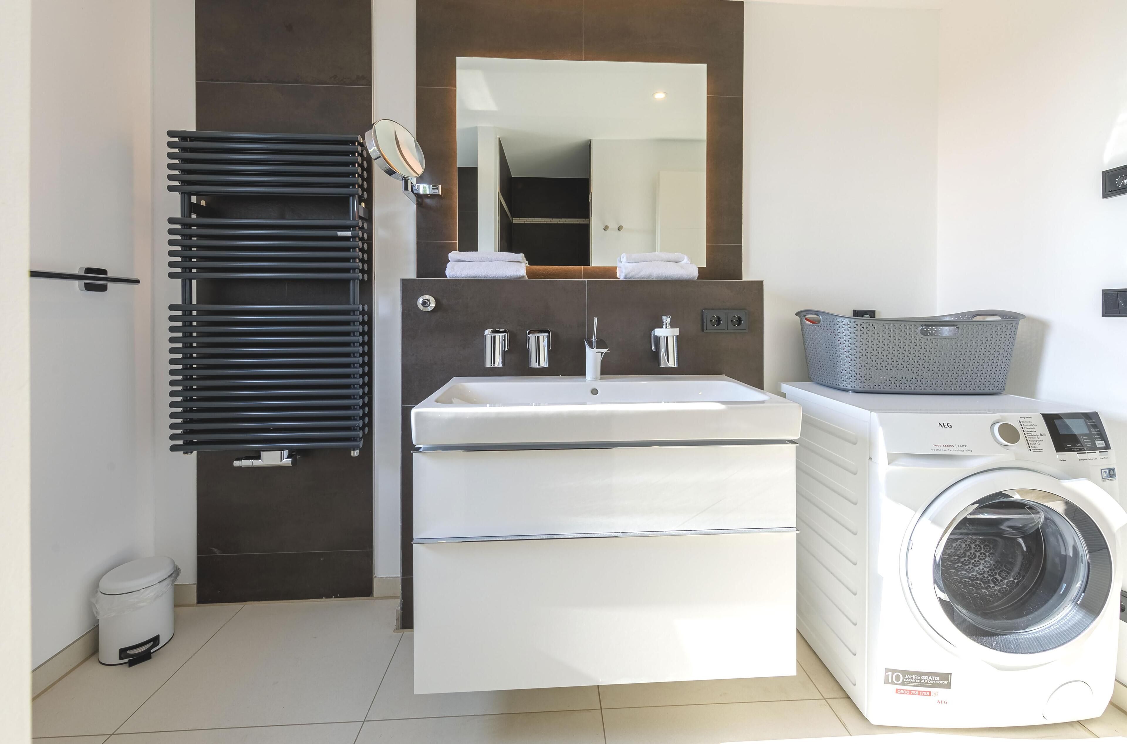 A modern bathroom with a white sink, a mirror, a black towel rack, a white washing machine, and a grey laundry basket.
