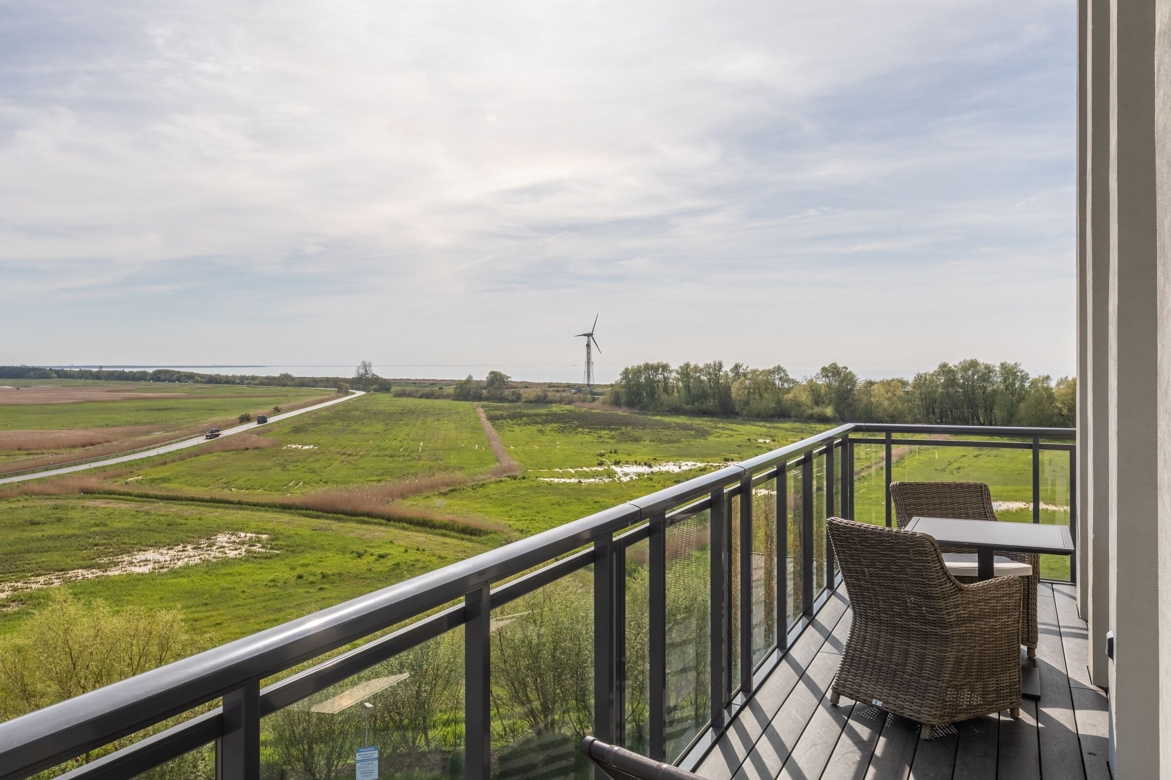 A balcony with wicker furniture, a view of a green landscape, and a wind turbine in the distance.