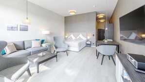 A modern hotel room with a sofa, two armchairs, a dining table, a bed with a headboard, and a TV mounted on the wall.